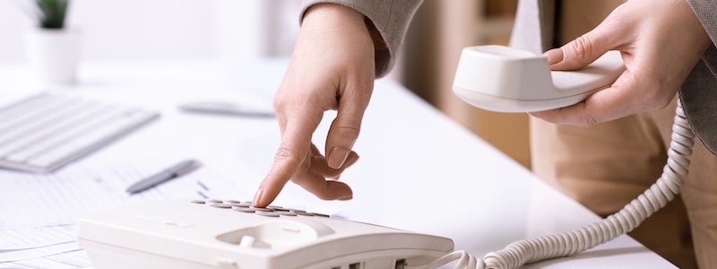 Eine Person hält den Hörer eines schnurgebundenen Telefons und wählt mit der anderen Hand eine Nummer auf der Tastatur.