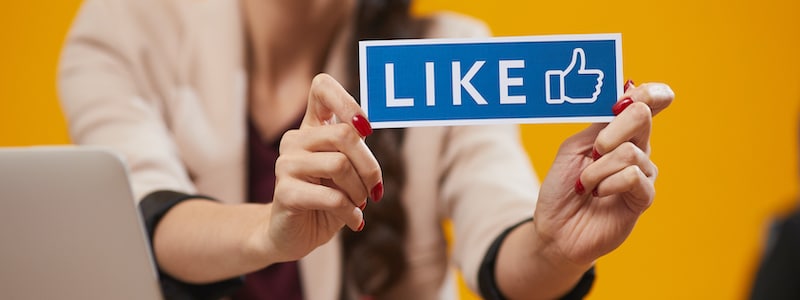 Person, die ein „Gefällt mir“-Zeichen mit einem Daumen-hoch-Symbol vor einem orangefarbenen Hintergrund hält, wobei links ein Teil eines Laptops sichtbar ist.