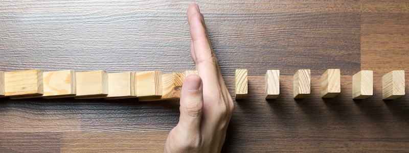 Eine Hand wird vertikal zwischen einer Reihe von Holzklötzen platziert und sorgt dafür, dass die Klötze nicht mehr nacheinander auf eine Holzoberfläche fallen.