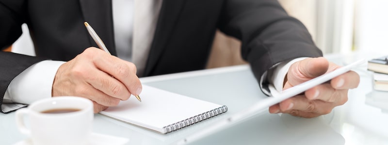 Eine Person im Anzug sitzt an einem Schreibtisch, schreibt mit einem Stift in einen Notizblock und hält ein Tablet in der Hand. Auf dem Schreibtisch steht eine Tasse Kaffee.
