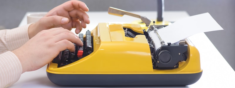 Eine Person tippt auf einer gelben Schreibmaschine, in die ein Blatt Papier eingelegt ist.