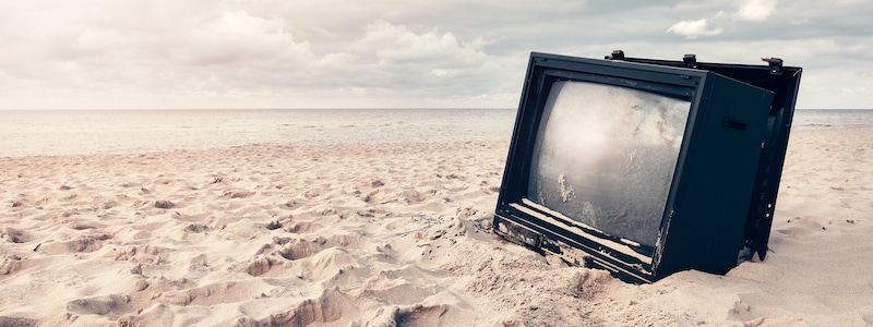 Ein kaputter Fernseher ist teilweise im Sand an einem Strand vergraben, im Hintergrund sind das Meer und ein bewölkter Himmel zu sehen.