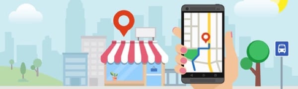 Hand hält ein Smartphone mit geöffneter GPS-Navigation, das eine Route zu einem Geschäft mit einer roten Standortmarkierung vor dem Hintergrund einer Stadtlandschaft mit Bäumen, Gebäuden und einer Bushaltestelle anzeigt.