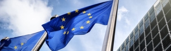 Vor einem modernen Glasgebäude wehen vor einem blauen Himmel Flaggen der Europäischen Union an Fahnenmasten.