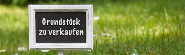Ein kleines Tafelschild auf einem grünen Rasen mit weißem Text auf Deutsch, auf dem „Grundstück zu verkaufen“ steht.
