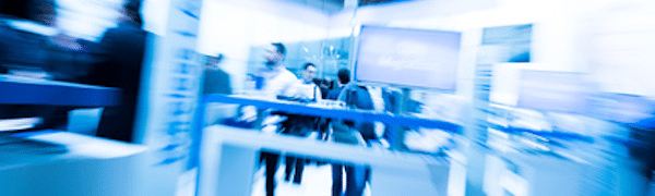 Unscharfes Bild von Menschen, die durch einen Ausstellungs- oder Messebereich mit bläulicher Beleuchtung und Displays gehen.