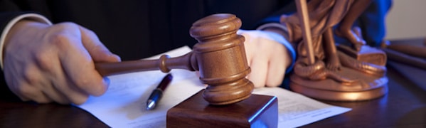 Ein Richter schlägt mit einem hölzernen Hammer auf einen Schreibtisch. Im Hintergrund sind juristische Dokumente und ein Stift zu sehen. Eine bronzene Statue der Justitia ist zu sehen.