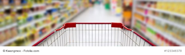 Im Vordergrund ist ein Einkaufswagen zu sehen, im Hintergrund sind die Gänge mit Lebensmitteln unscharf dargestellt.