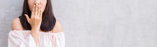 Eine Person mit langen dunklen Haaren und einem schulterfreien Oberteil bedeckt den Mund mit der Hand, während sie vor einer grau strukturierten Wand steht.