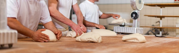 Drei Bäcker in weißen Hemden kneten und formen Teig auf einer Holztheke in einer Küche mit Waagen und Regalen.