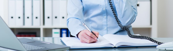 Person in einem blauen Hemd schreibt in ein Notizbuch, während sie mit einem schnurgebundenen Telefon telefoniert, im Hintergrund sind ein Laptop und Ordner zu sehen.