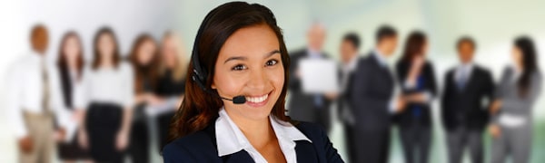 Eine Frau mit Headset lächelt in die Kamera, während sie vor einer unscharfen Büroumgebung mit mehreren Personen in Business-Kleidung steht.