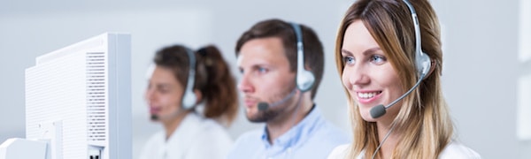 Drei Kundendienstmitarbeiter mit Headsets arbeiten an Computerstationen in einem hell erleuchteten Büro. Im Vordergrund steht eine lächelnde Frau.