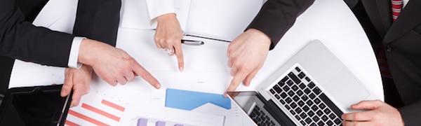 Vier Hände zeigen auf unterschiedliche Abschnitte verschiedener Diagramme und Grafiken, die auf einem Tisch ausgebreitet sind. In der Nähe steht ein Laptop.