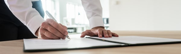 Person im weißen Hemd unterschreibt in einer hellen Büroumgebung ein Dokument auf einem Schreibtisch.