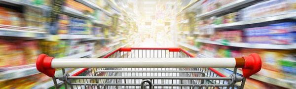 In einem hell erleuchteten Lebensmittelladen wird ein Einkaufswagen durch einen Gang geschoben. Auf beiden Seiten sind Regale mit verschiedenen Produkten gefüllt.