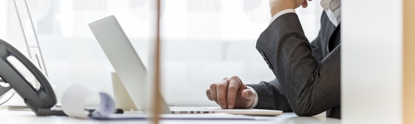 Eine Person im Anzug sitzt an einem Schreibtisch und arbeitet an einem Laptop. Auf dem Schreibtisch sind ein Festnetztelefon, Dokumente und ein Stift zu sehen.
