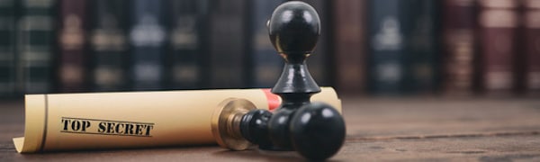 Ein zusammengerolltes Dokument mit der Aufschrift „Streng geheim“ und einem schwarzen Stempel liegt auf einer Holzoberfläche vor dem unscharfen Hintergrund eines Bücherregals.