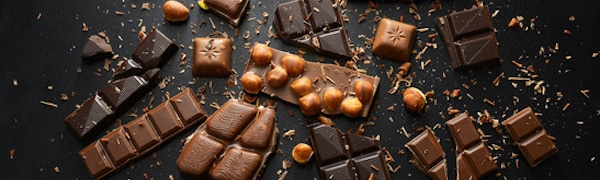 Verschiedene Stücke dunkler und Vollmilchschokolade, einige davon mit Haselnüssen, auf einer dunklen Oberfläche ausgelegt und mit Schokoladenraspeln darüber verstreut.