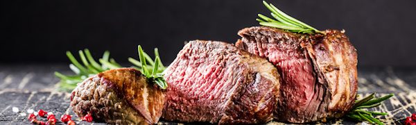 Drei Scheiben medium gebratenes Steak, garniert mit Rosmarin auf einer Holzoberfläche vor dunklem Hintergrund.