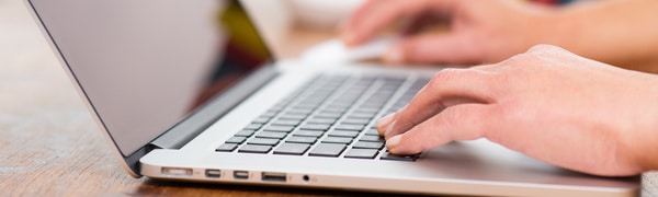 Nahaufnahme von Händen, die auf einer Laptoptastatur tippen. Der Laptop steht auf einer Holzoberfläche.