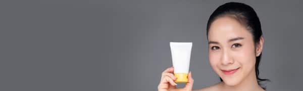 Eine Person mit zurückgebundenem dunklem Haar lächelt und hält vor einem schlichten, grauen Hintergrund eine weiß-gelbe Tube mit einem Hautpflegeprodukt.