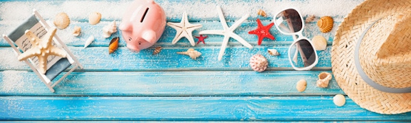 Auf einer blauen Holzoberfläche sind Gegenstände zum Thema Strand angeordnet, darunter ein Strandstuhl, ein Sparschwein, Seesterne, Muscheln, eine Sonnenbrille und ein Strohhut.