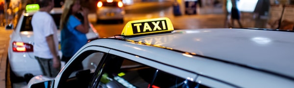 Nahaufnahme eines geparkten Taxis mit einem leuchtenden „TAXI“-Schild auf dem Dach, mit Menschen und einem weiteren Auto im Hintergrund auf einer Stadtstraße bei Nacht.