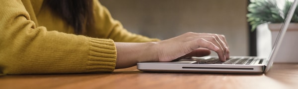 Eine Person in einem gelben Pullover tippt an einem Holzschreibtisch auf einem Laptop. Im Hintergrund ist eine Pflanze zu sehen.