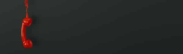 Auf der linken Seite des Bildes hängt vor einem dunklen Hintergrund ein roter schnurgebundener Telefonhörer.