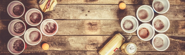 Draufsicht auf einen Holztisch mit mehreren für Bierpong angeordneten roten Plastikbechern, zwei Tischtennisbällen und Bierdosen.