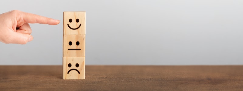 Eine Hand zeigt auf einen Stapel von drei Holzklötzen mit Gesichtern. Der obere Klotz hat ein fröhliches Gesicht, der mittlere ein neutrales Gesicht und der untere ein trauriges Gesicht.