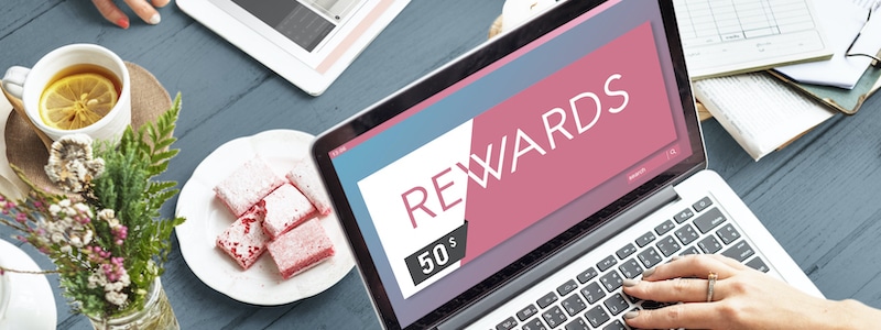 Ein Laptop mit einem Bildschirm und dem Text „BELOHNUNGEN“ auf einem Tisch neben einer Tasse Tee, einem Teller mit rosa Gebäck, einem Blumenstrauß und einem offenen Notizbuch mit einem Stift.