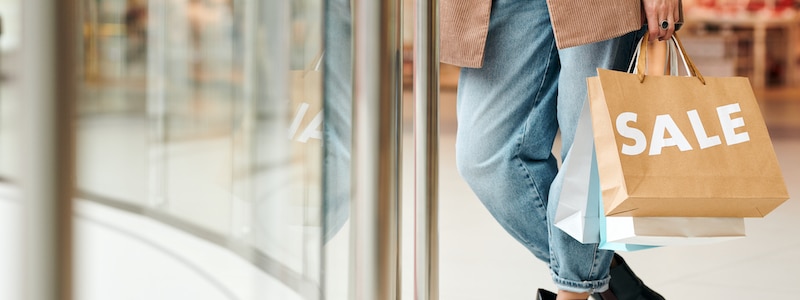 Person trägt Einkaufstüten mit dem Etikett „SALE“ und geht an einem Schaufenster vorbei.