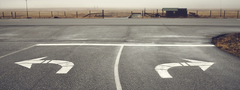 Eine Straße mit aufgemalten Pfeilen, die Links- und Rechtsabbiegen anzeigen, führt zu einer leeren Autobahn unter einem bewölkten Himmel. Im Hintergrund sind ein Zaun und ein kleines Gebäude zu sehen.