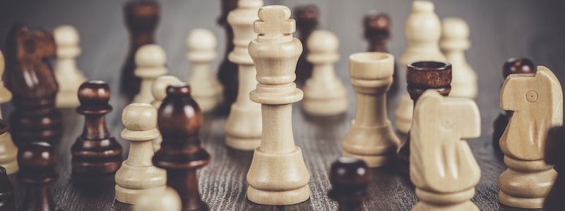 Eine Nahaufnahme verschiedener Schachfiguren aus Holz, die auf einer Holzoberfläche verstreut sind, mit einer weißen Königsfigur in der Mitte. Die Figuren bestehen aus einer Mischung aus hellem und dunklem Holz.