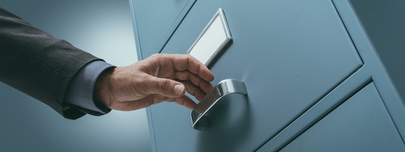 Eine Hand greift in einer Büroumgebung nach dem Griff einer blauen Aktenschrankschublade.