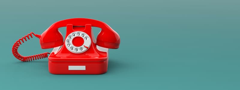 Auf einer grünen Fläche vor grünem Hintergrund steht ein rotes Telefon mit Wählscheibe und Spiralkabel.