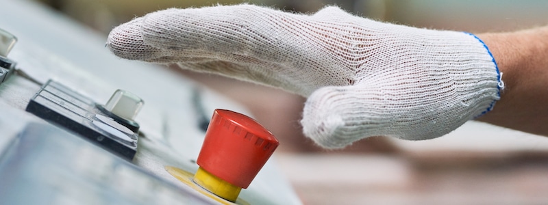Eine mit einem weißen Handschuh bekleidete Hand schwebt über einem roten Not-Aus-Schalter auf einem Bedienfeld.
