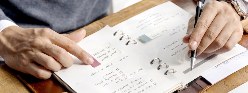 Eine Person verwendet einen Stift, um in einen Ringbuchplaner zu schreiben, während sie mit der anderen Hand auf einen Abschnitt der Seite zeigt.