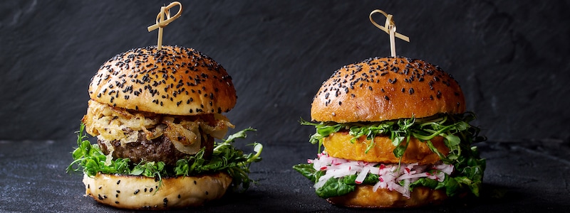 Zwei Gourmetburger auf einer dunklen Oberfläche. Der linke Burger hat karamellisierte Zwiebeln und Grünzeug, der rechte Burger hat Radieschenscheiben und Grünzeug. Beide haben Sesambrötchen, die mit Holzspießen gehalten werden.