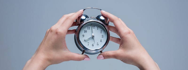 Ein Paar Zeiger, die einen silbernen analogen Wecker vor einem schlichten grauen Hintergrund halten. Die Uhr zeigt kurz nach 10:10 Uhr an.