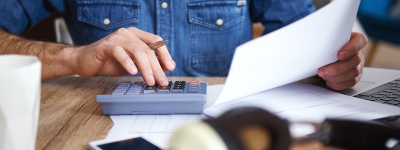 Person, die einen Taschenrechner benutzt und ein Blatt Papier hält; sie arbeitet an einem Schreibtisch mit Dokumenten, einem Smartphone, Kopfhörern und einem Laptop.