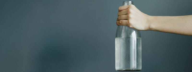 Eine Hand hält vor einem schlichten grauen Hintergrund eine klare Glasflasche, die zur Hälfte mit Wasser gefüllt ist.