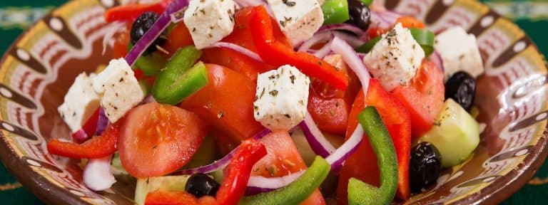 Ein bunter Salat mit Fetakäsewürfeln, roten und grünen Paprika, roten Zwiebelscheiben, Tomaten, schwarzen Oliven und Gurken in einer gemusterten Schüssel.